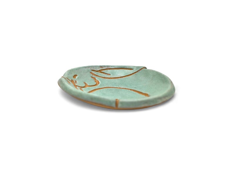 May include: A small, round, ceramic dish with a light teal glaze. The dish features a brown, abstract design on the surface. The rim of the dish has a slightly raised edge. The dish is likely handmade and suitable for holding small items.