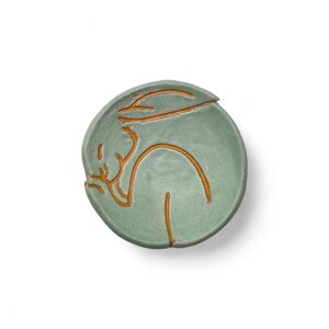 May include: A round, light green ceramic dish with a stylized, golden-brown outline of a person. The person is depicted in a reclining pose. The dish has a lid with a small, curved handle. The design is minimalist and artistic.