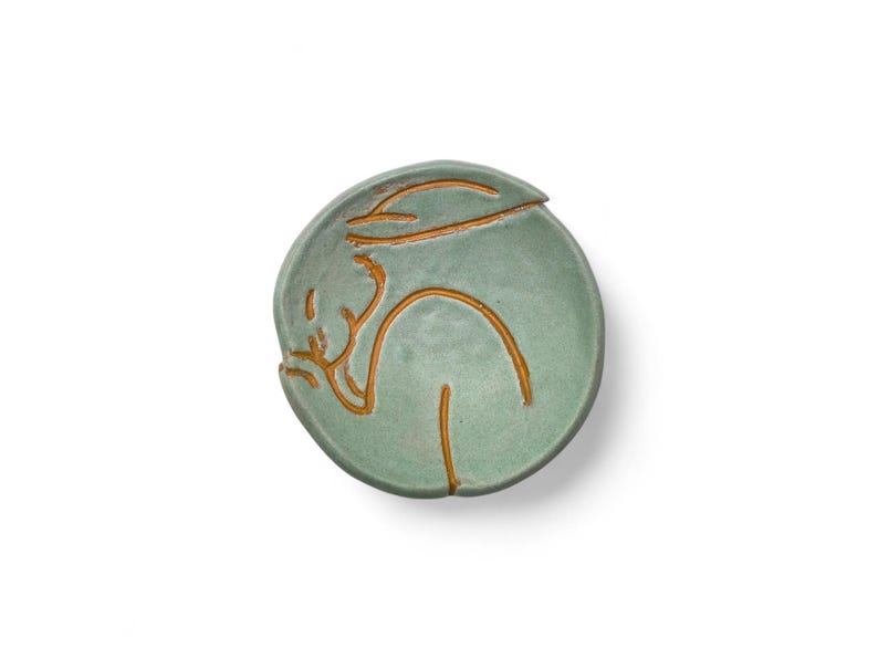 May include: A round, light green ceramic dish with a stylized, golden-brown outline of a person. The person is depicted in a reclining pose. The dish has a lid with a small, curved handle. The design is minimalist and artistic.