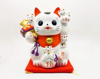 Puede incluir: Figura de cerámica blanca Maneki-Neko, o gato de la suerte, con la pata levantada. El gato sostiene una moneda de oro y una bolsa roja y blanca. El gato está rodeado de gatos blancos más pequeños. El gato está sentado en un cojín rojo con la palabra "福" (que significa "buena suerte") escrita en él.