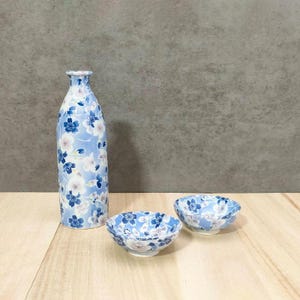 May include: A set of three ceramic sake serving pieces with a blue and white floral pattern. The set includes a bottle and two small cups.