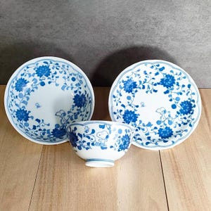 May include: A three-piece ceramic dish set featuring a blue floral pattern on a white background. The set includes two plates and a bowl. The design incorporates a cartoon character. The dishes are arranged on a wooden surface.