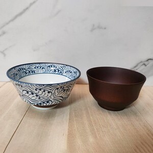 May include: Two bowls, one white with a blue wave pattern and one brown. Both bowls are round and have a slight lip.
