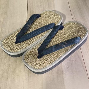 May include: A pair of traditional Japanese geta sandals. The sandals feature a woven straw footbed, a dark blue fabric thong, and a gray sole. The thong has a subtle floral pattern. The sandals are designed for comfort and style.