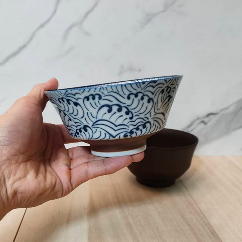 May include: A white ceramic bowl with a blue and white wave pattern. The bowl is being held in a hand.