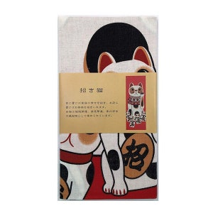 May include: A white cotton tea towel with a black and white illustration of a Japanese Maneki-Neko, or beckoning cat. The cat is sitting on a red and gold mat with the Japanese character for "fortune" on it. The text on the label reads "招き猫" which translates to "beckoning cat" in English. The text on the label also reads "家に置けば家族の幸せを招き、お店に受けばお客様を招きいれます。自揚は福後開道、京売繁盛、家内安全の縁起物として高められています。"
