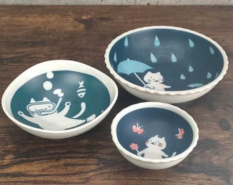May include: Set of three blue and white ceramic bowls with cat illustrations. The bowls feature a cat scuba diving, a cat under an umbrella in the rain, and a cat with butterflies.