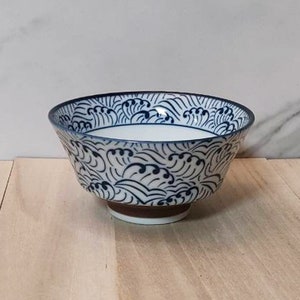 May include: A white ceramic bowl with a blue and white wave pattern. The bowl has a rounded shape and a wide rim.