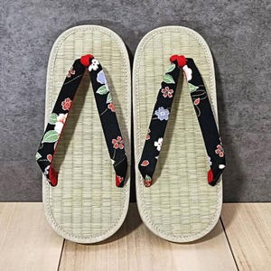 May include: Pair of traditional Japanese geta sandals. The sandals have a light beige woven platform with black straps. The straps are decorated with a floral pattern in red, white, and purple. The thong is red.