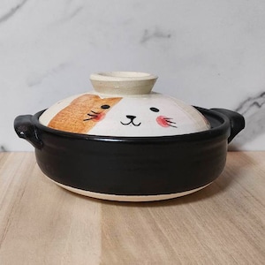 May include: A black and white ceramic pot with a cat face design on the lid. The lid is off and the pot is sitting on a wooden surface.