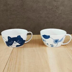 以下が含まれることがあります： 猫のイラストが描かれた白いセラミックマグカップが2つ。1つのマグカップには濃い青色の猫のデザイン、もう1つには青と白の猫の顔が描かれています。どちらのマグカップにも取っ手があり、木製の表面に置かれています。