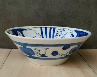 May include: A white ceramic bowl featuring a blue and white cat design. The bowl is decorated with cats in various poses, including a striped cat, a spotted cat, and a cat with its tail curled around its body.
