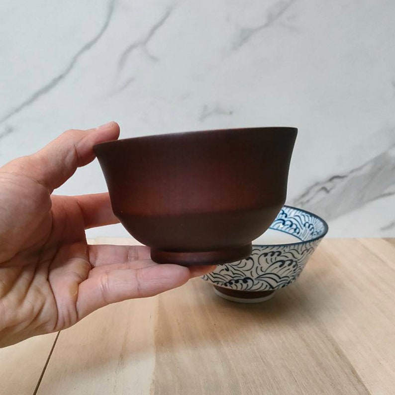 May include: A dark brown ceramic bowl with a slightly flared rim. The bowl is held in a hand, and a smaller blue and white patterned bowl is visible in the background.