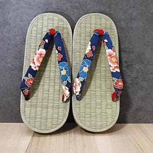 May include: Pair of traditional Japanese geta sandals. The sandals have a light beige woven platform and navy blue straps adorned with pink and red floral patterns. The straps are accented with red fabric at the ends.