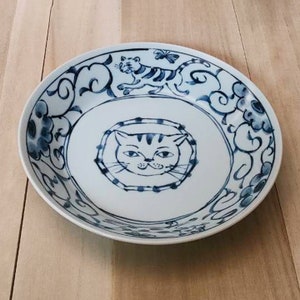 May include: A white ceramic plate with a blue and white design featuring a cat and a tiger. The plate has a blue and white floral pattern around the edge.