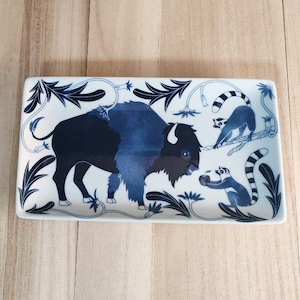 May include: A white rectangular ceramic tray with a blue and white design. The tray features a blue bison, two blue and white lemurs, and a blue floral pattern.