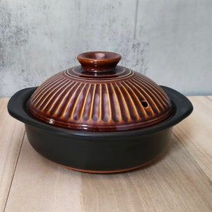 以下が含まれることがあります： 蓋付きの茶色と黒の陶器製の鍋。鍋はリブ模様で、蓋に小さな穴が開いています。鍋は木製の表面の上に置かれています。