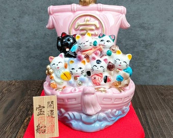 以下が含まれることがあります： 7匹の猫が乗った船の形をしたピンクの陶器製の招き猫の置物。猫はそれぞれ色が異なり、表情も違います。船は金貨と赤白の旗で飾られています。船の前方には「幸福」の文字が書かれています。