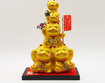 以下が含まれることがあります： 赤いアクセントと日本語の文字が書かれた、金の招き猫の置物が5体積み重ねられています。猫はピラミッド型に積み重ねられており、一番小さい猫が上に、一番大きい猫が下にあります。一番下の猫は、「幸せ」と「連れて」と書かれた旗を持っています。