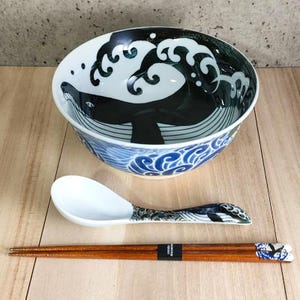 May include: A set of Japanese-style tableware featuring a blue and white ceramic bowl with a black whale design, a white ceramic spoon with a black whale design on the handle, and a pair of wooden chopsticks with a black whale design on the ends.