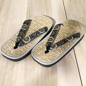 May include: A pair of traditional Japanese geta sandals. The sandals feature a woven straw footbed, a black sole, and a strap with a floral pattern in shades of blue, black, and white. The edges are trimmed with a light gray border.
