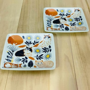 May include: Two white ceramic plates with a cat and flower design. The plates feature orange, black, and white cats with a floral pattern in shades of white, green, and yellow.