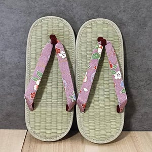 May include: A pair of traditional Japanese geta sandals. The sandals feature a light green woven straw base with a rounded toe and a purple fabric strap. The straps are decorated with a floral pattern in white, red, and green.
