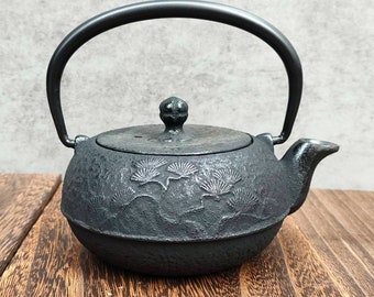Könnte beinhalten: Schwarze gusseiserne Teekanne mit einem Kiefernbaumdesign. Die Teekanne hat einen runden Körper und einen gebogenen Griff.