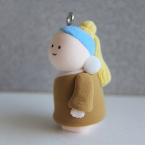 May include: A handmade figurine of a woman with a pearl earring, inspired by the famous painting. The figure has a tan face, a blue headscarf, and a yellow bun. The woman is wearing a brown dress and a white pearl earring.