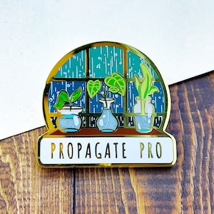 Cute enamel pin of a rainy windowsill filled with little jars of propagating plants. The text reads Propagate Pro. This gardener accessory is discounted due to a flaw that can be seen in the enamel, but its sale price doesnt reduce its beauty!