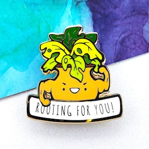 May include: A yellow and green enamel pin of a potted plant with a smiling face and flexing arms. The plant is in a yellow pot and has green leaves. The pin has a white banner that says "ROOTING FOR YOU!"