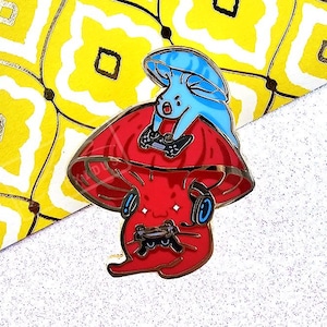 Op de afbeelding: Een rode en blauwe, paddenstoelvormige emaille pin met twee cartoonfiguren erin. De figuren dragen koptelefoons en houden videogamecontrollers vast.