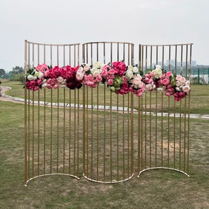 May include: A gold metal backdrop with a curved design, decorated with pink, white, and green artificial flowers. The backdrop is made of thin metal rods and is perfect for creating a beautiful and elegant background for photos or events.