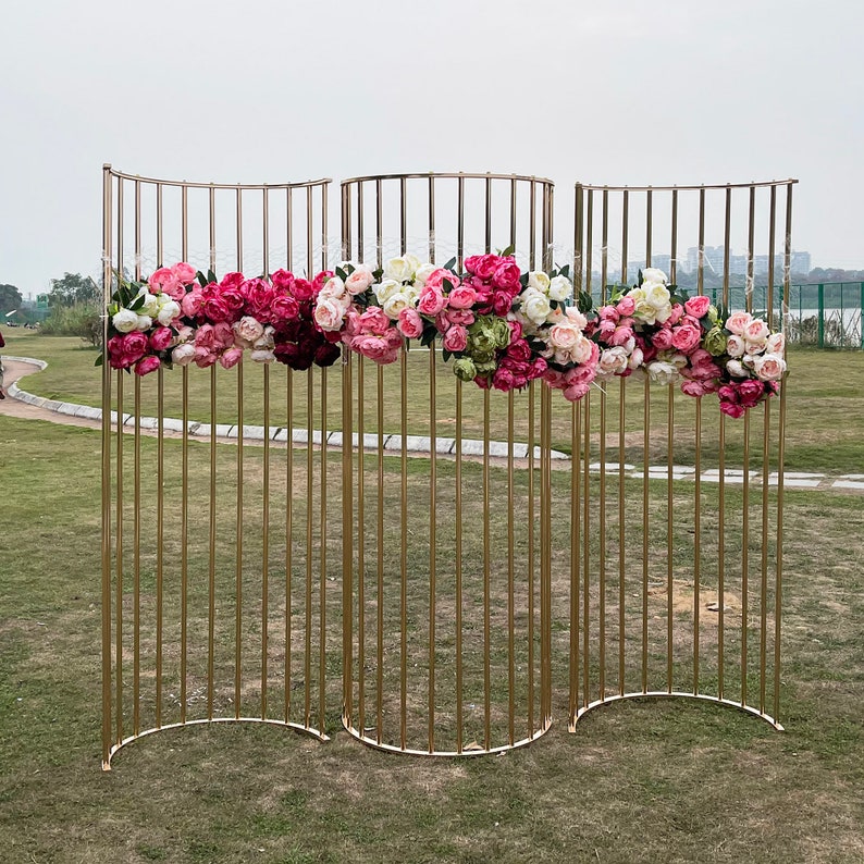 May include: A gold metal backdrop with a curved design, decorated with pink, white, and green artificial flowers. The backdrop is made of thin metal rods and is perfect for creating a beautiful and elegant background for photos or events.