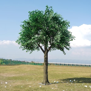 Op de afbeelding: Een groene kunstmatige boom met een bruine stam, staande op een grasveld met een brug op de achtergrond.