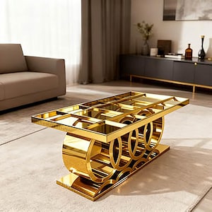 Op de afbeelding: Een rechthoekige salontafel met een reflecterend goudkleurig metalen frame met een rasterpatroon aan de bovenkant en een basis bestaande uit een reeks onderling verbonden cirkelvormige metalen ringen. De tafel heeft een helder glazen blad.