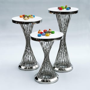 May include: Three white round tables with silver, twisted wire bases. The tables are stacked on top of each other, with the largest table at the bottom and the smallest table at the top. Each table has a variety of colorful macarons and pastries on top.