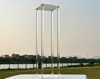 May include: A clear acrylic pedestal with four legs and a square top. The pedestal is standing on a white tablecloth.