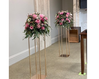 May include: Two gold metal stands with pink and white flower arrangements. The stands are tall and slender with a square base.