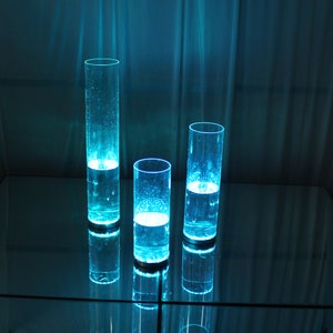 May include: Three clear glass cylinders filled with blue liquid, illuminated from within, creating a glowing effect. The cylinders are arranged on a reflective surface.