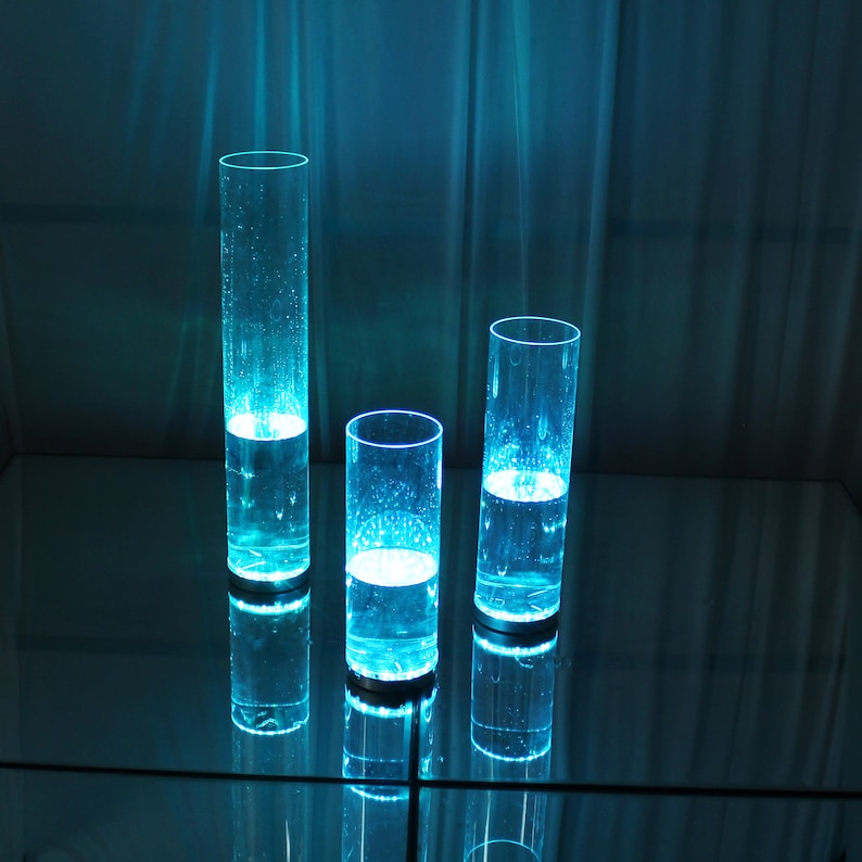 May include: Three clear glass cylinders filled with blue liquid, illuminated from within, creating a glowing effect. The cylinders are arranged on a reflective surface.
