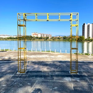 May include: A golden metal archway structure, ideal for outdoor events. The arch features a rectangular frame design with multiple shelves. The structure is set against a backdrop of a lake, trees, and buildings under a clear blue sky.