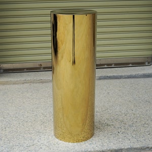 Puede incluir: Un pedestal cil&iacute;ndrico dorado y alto. El pedestal tiene una superficie lisa y reflectante y una parte superior plana. La base tambi&eacute;n es plana. El pedestal est&aacute; hecho de un material met&aacute;lico brillante. El fondo es de color neutro.