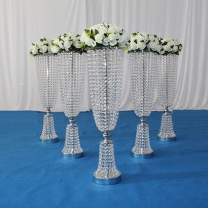 May include: Five silver crystal wedding centerpieces with white flowers. Each centerpiece has a tall, slender vase with a crystal base and a crystal chain hanging from the top.