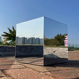 May include: A large, silver, mirrored cube reflecting the surrounding environment. The cube is sitting on a brick patio.