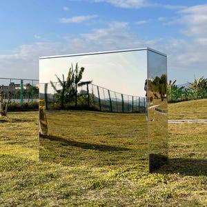 May include: A large, rectangular, silver-colored, mirrored sculpture stands on a grassy lawn. The sculpture reflects the surrounding landscape, including trees, a fence, and a blue sky.