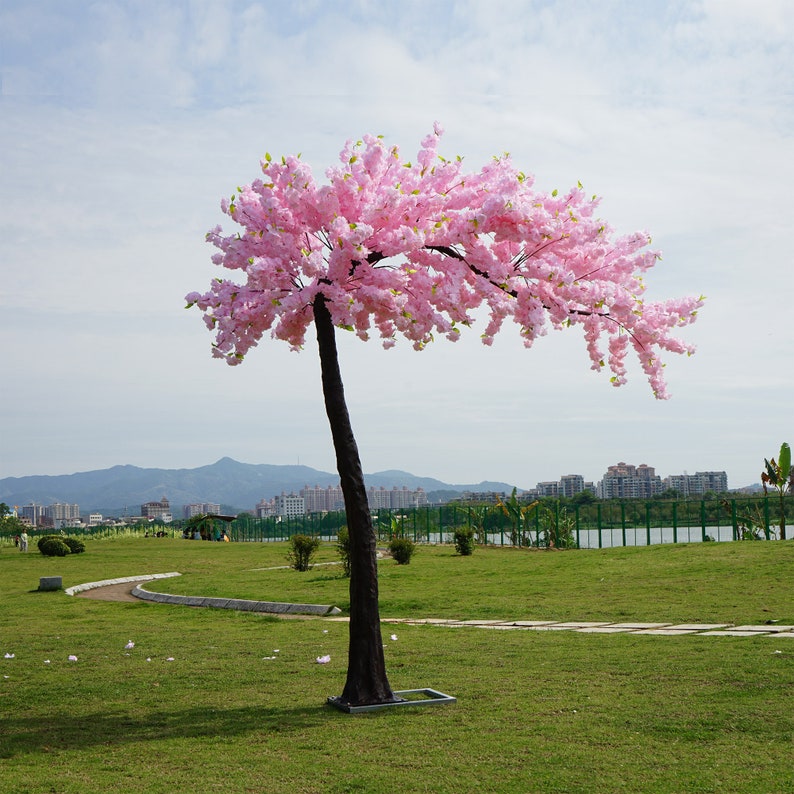 May include: A pink artificial cherry blossom tree with a brown trunk and branches. The tree is in a grassy field with a city skyline in the background.