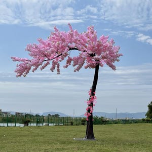 May include: A pink artificial cherry blossom tree with a brown trunk. The tree is standing on a green grassy lawn with a blue sky in the background.