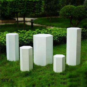May include: A collection of five white, octagonal pedestals of different heights, positioned on a lawn. The pedestals are geometric in design. The text "SET A" is visible in the lower left corner.