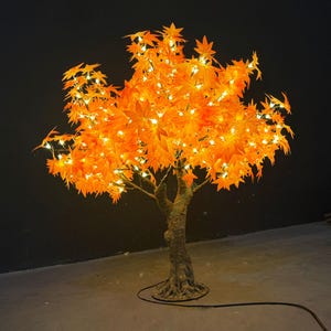 Könnte beinhalten: Ein beleuchteter künstlicher Ahornbaum mit leuchtend orangefarbenen Blättern. Der Baum ist mit kleinen, warmweißen Lichtern geschmückt, die einen leuchtenden Effekt erzeugen. Der Stamm ist strukturiert, und der Baum steht vor einem dunklen Hintergrund.